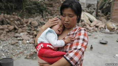 四川雅安蘆山縣龍門鄉一名倖存婦女抱着孩子站在已經倒塌的房子旁（21/4/2013）