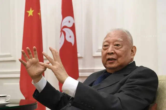 董建华坐在沙发上,背景是中国国旗和香港区旗