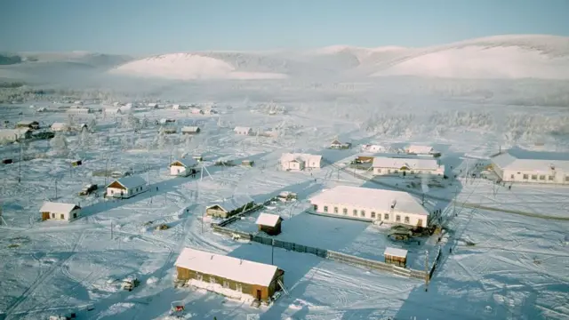 一座村庄的鸟瞰图,数座低矮建筑散布在平坦的雪原上,后方可见白雪覆盖的山脉。