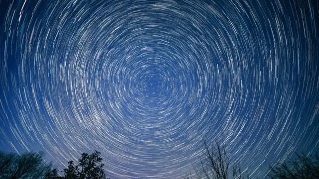 一张夜空长时间曝光照片,显示星轨围绕着一个中心点呈圆形运动;天空呈中等蓝色,多棵树木的树梢沿着画面底部可见。